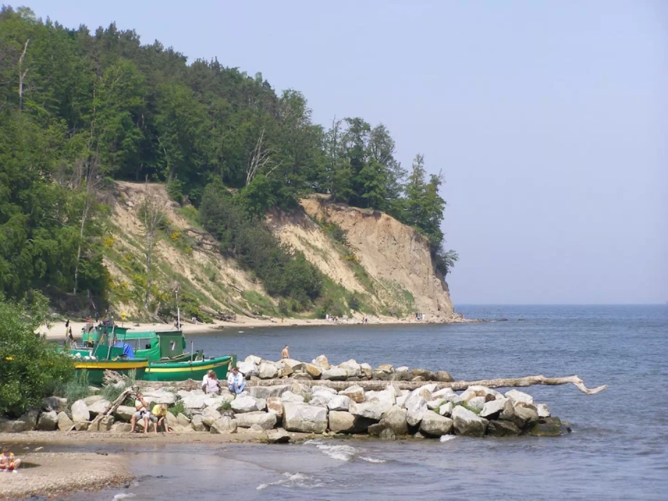Zdjęcie przedstawiające zatokę Gdańską i Klif Orłowski. Na pierwszym planie falochron, zielona łódka rybacka oraz grupa turystów wypoczywających na słońcu