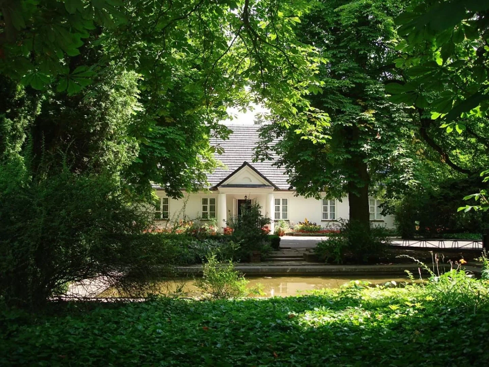 Budynek dworku w Żelazowej Woli. Minimalistyczny budynek z białą elewacją, kolumnadą przed wejściem i czarnym dachem. Przed nim znajduje się park pełen drzew i krzewów, a także staw.